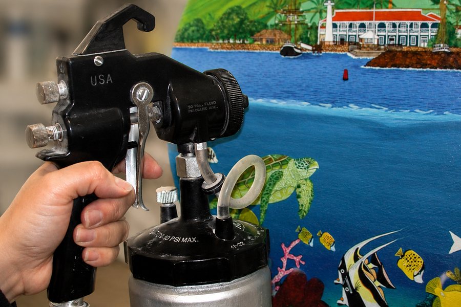 A spray gun applying protective varnish to a painted harbour scene.